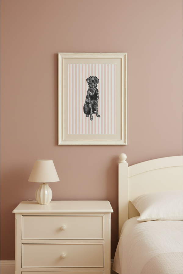 A framed vertical art print featuring a black dog illustration on a cream striped background, hanging on a dusty pink bedroom wall above a white nightstand and bed.