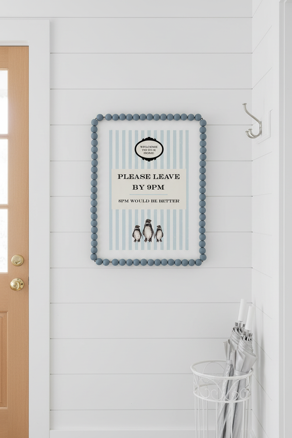 A framed decorative wall sign with a blue beaded border hangs on a white shiplap wall in a hallway. The sign has a cream center with light blue vertical stripes, black text reading 'PLEASE LEAVE BY 9PM', and small penguin illustrations near the bottom.