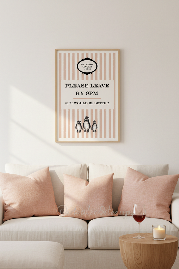 A framed vertical art print with pink and cream stripes and penguin illustrations hangs on a white wall above a beige sofa with blush pink pillows. A small wooden side table with a glass of red wine and a candle sits in the foreground.