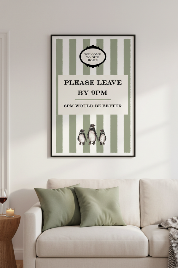 A framed vertical wall art print with green and white stripes, black text reading 'PLEASE LEAVE BY 9PM', and three penguin illustrations, displayed above a light beige sofa in a styled interior.