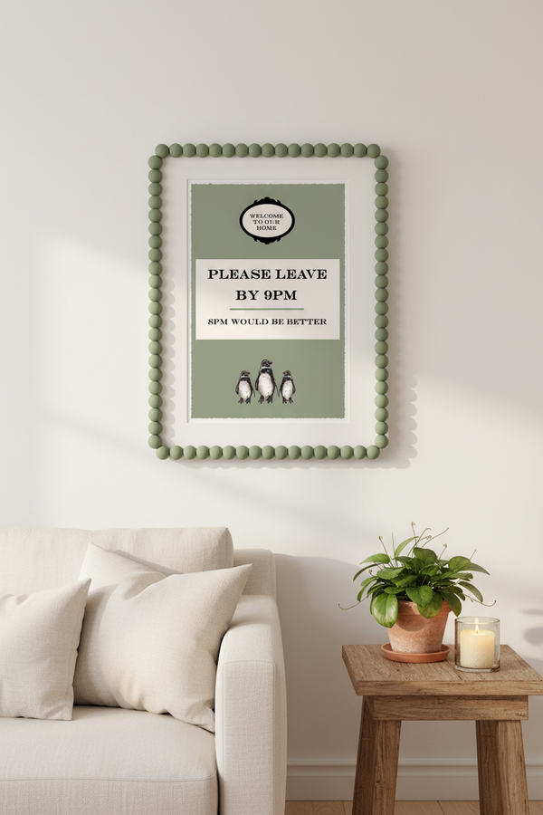 Please Leave By 9pm art print in a sage green bobble frame, hung on a white wall above a cream sofa and wooden side table with a plant and candle, in a bright minimal living room.