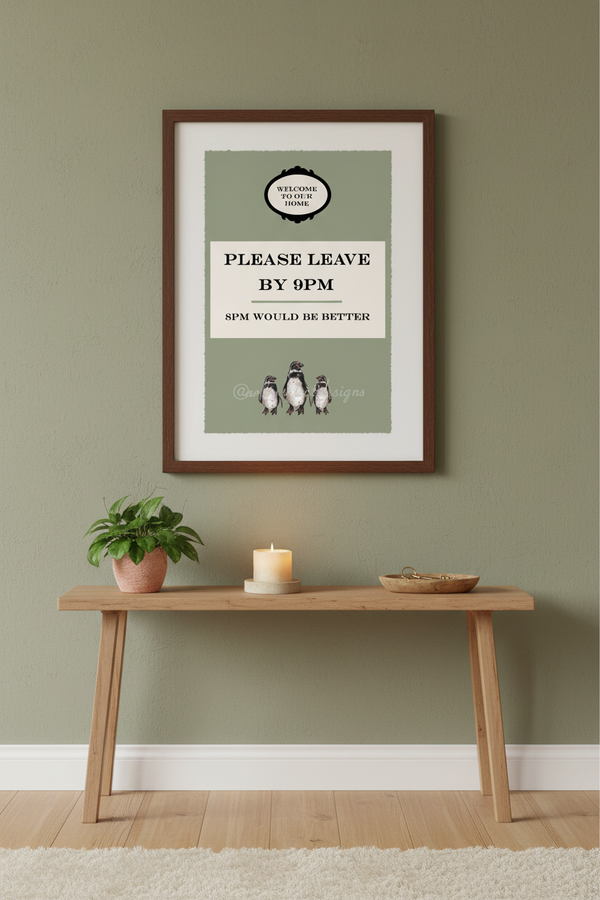 A framed art print featuring a vintage-style 'Please Leave By 9pm' sign with penguin illustrations, displayed on a sage green wall above a wooden console table with a plant and candle.