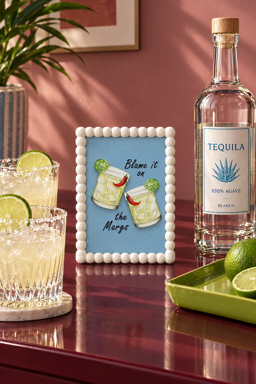 Blame It on the Margs A5 print in a white bobble frame on a burgundy home-bar surface, styled with cut-glass tumblers, a tequila bottle and a lime-green tray against a muted rose wall.