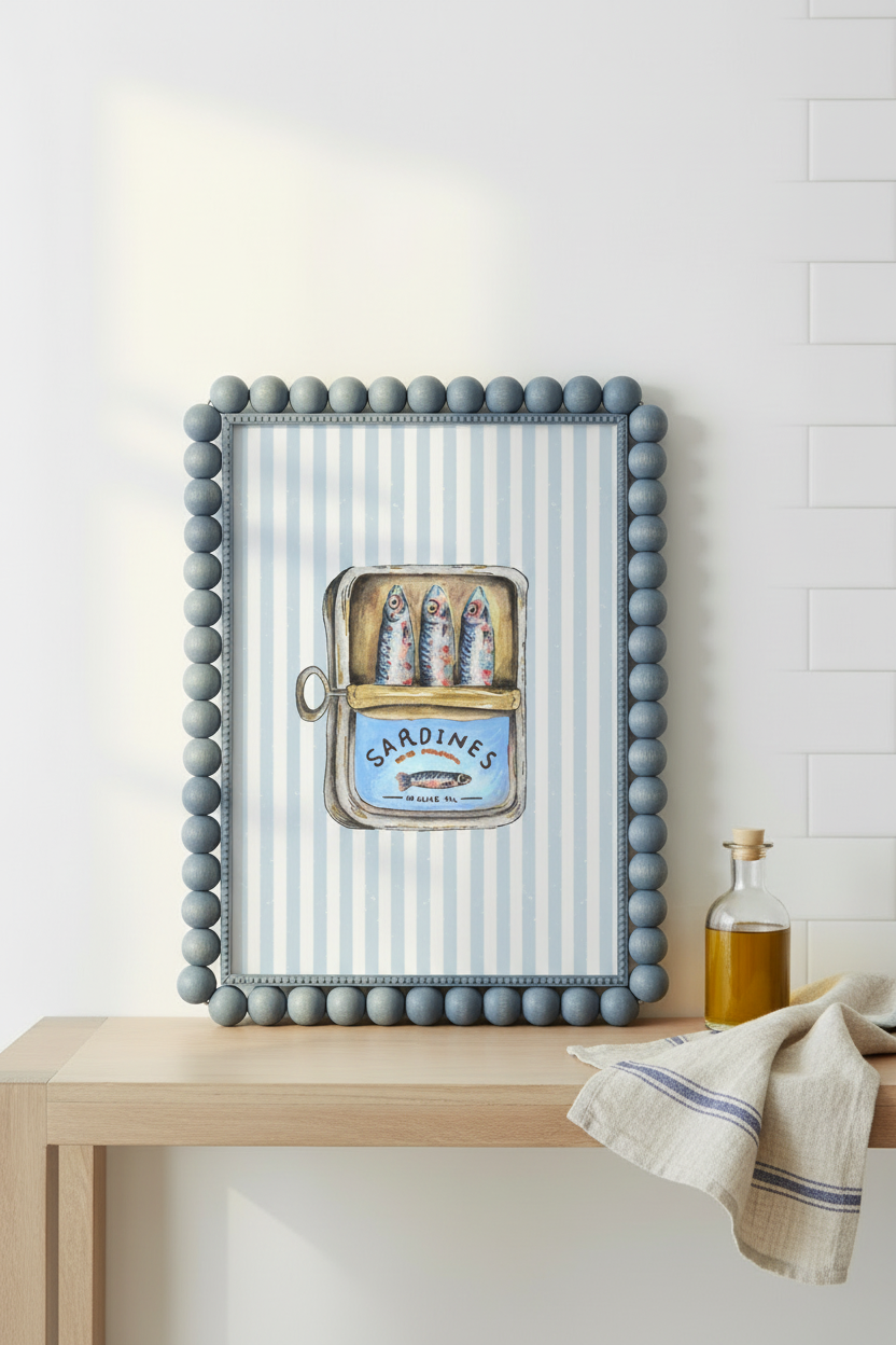 A framed decorative wall piece with a blue beaded border and pale blue-and-white vertical stripes, featuring an illustration of sardines in a can labeled Sardines. It is displayed on a light wood table against a white wall, with a small bottle and folded cloth nearby.