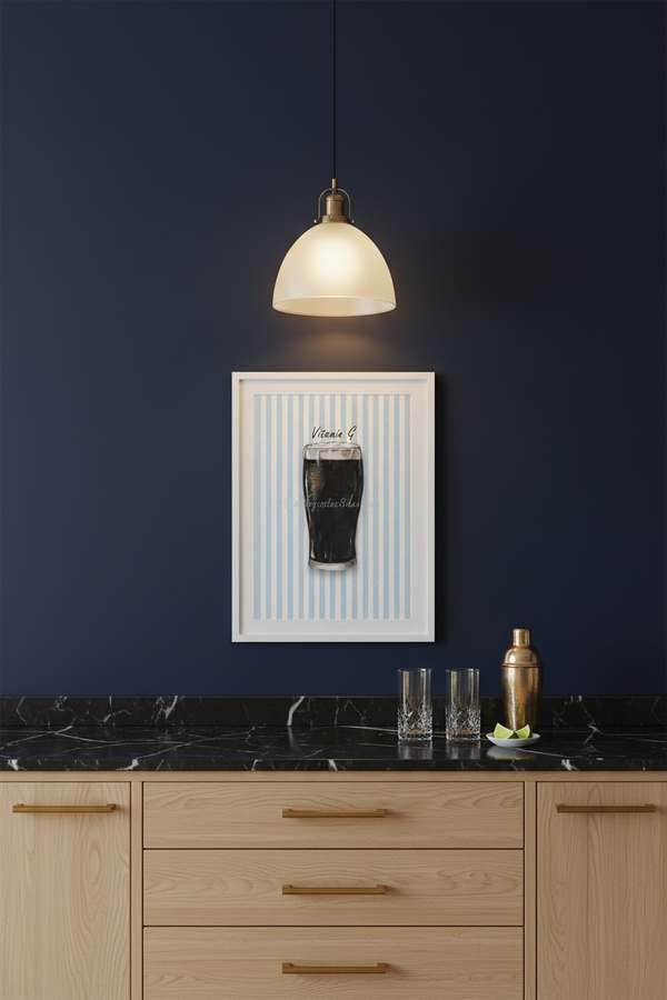 A framed wall art print featuring a dark beer glass on a light blue striped background, hanging on a dark navy wall above a wooden counter with a hanging pendant light.